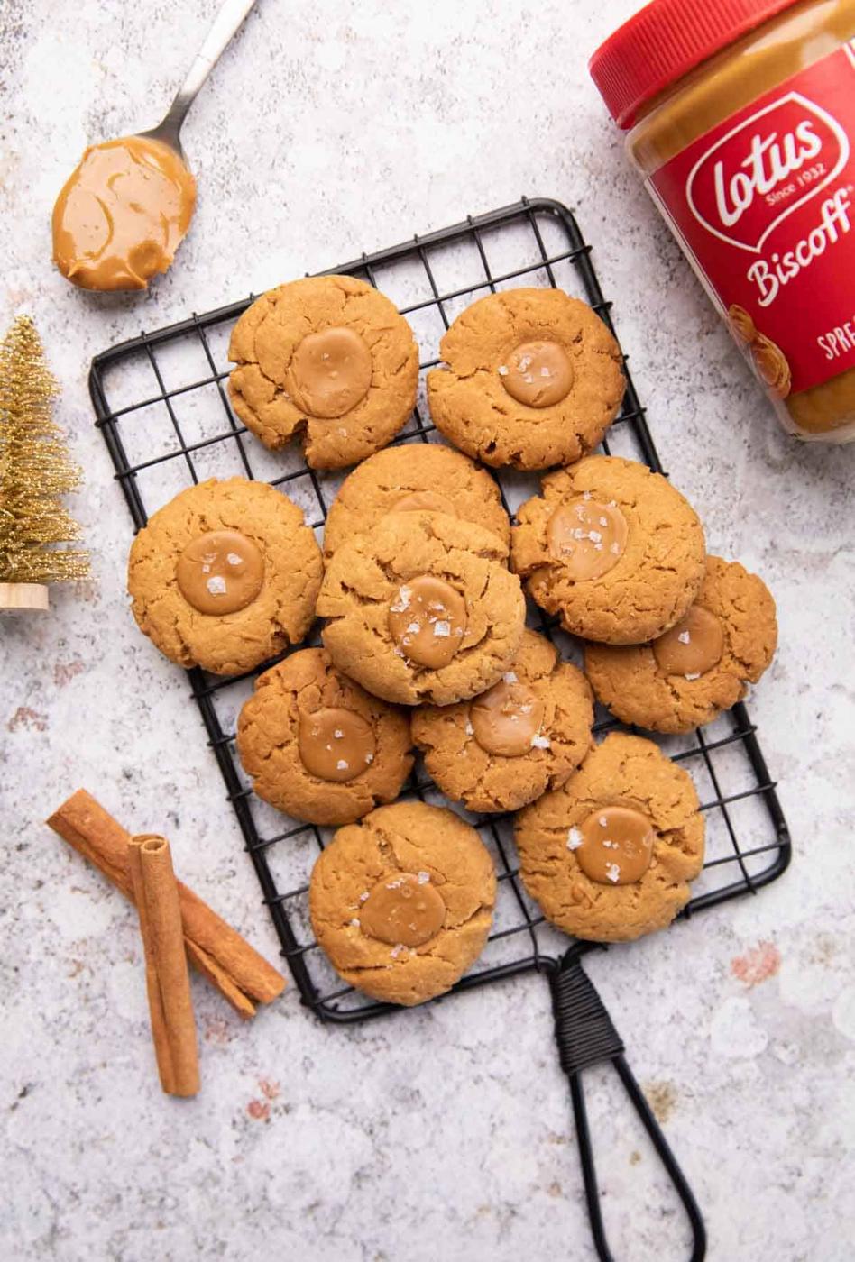 A shot of multiple biscoff thumbprint cookies stacked one on top of another on a black grill along with a spoon of biscoff, a golden christmas tree, two cinnamon sticks and a jar of biscoff on the sides of the grill.