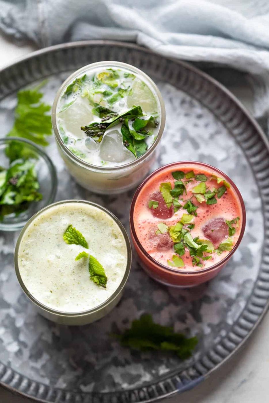 Three glasses of chaas served on a metal plate with curry leaves and coriander on the side