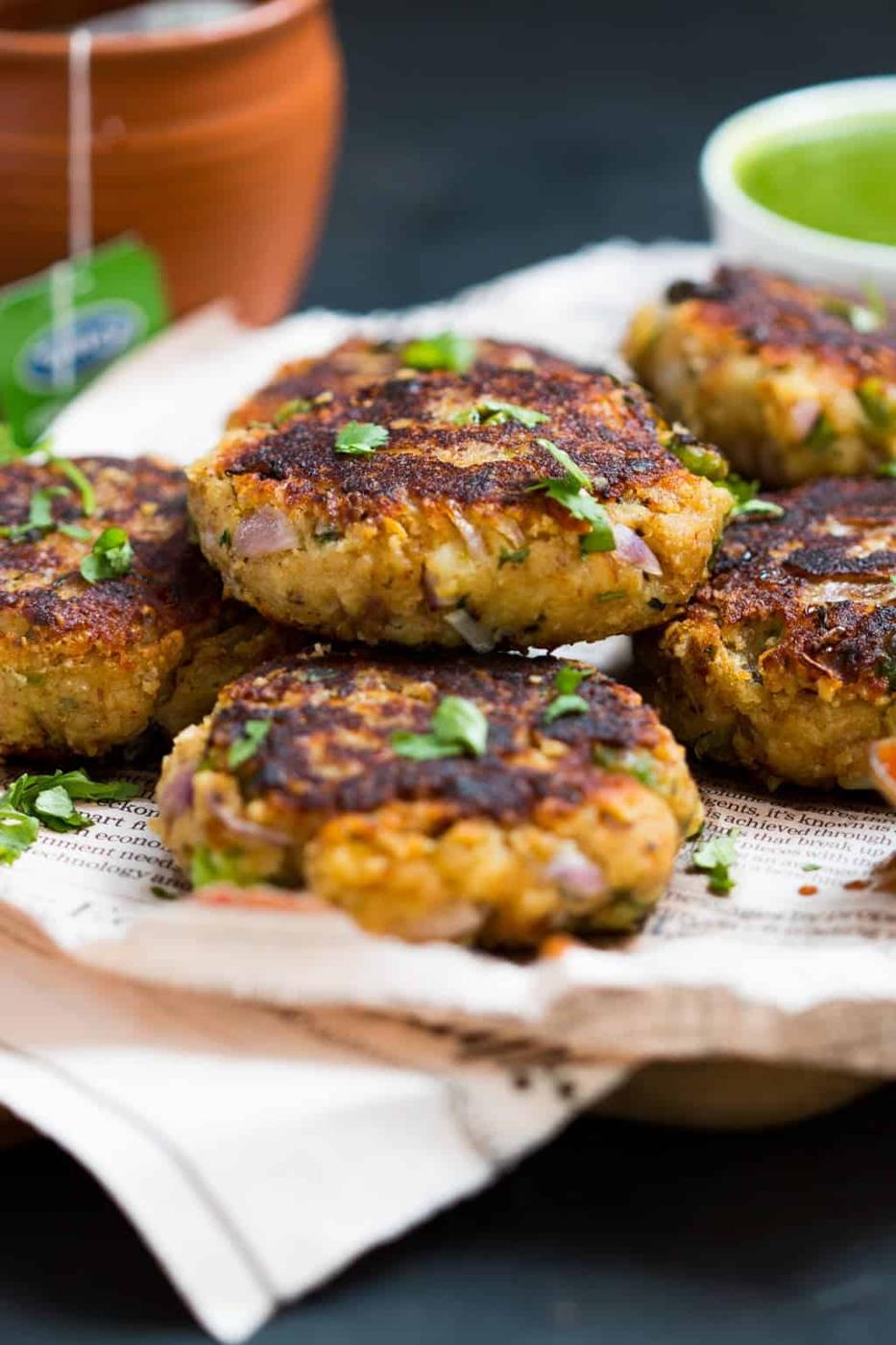 aloo paneer tikkis stacked on a brown board with newspaper alongside a small of chutney and some tea