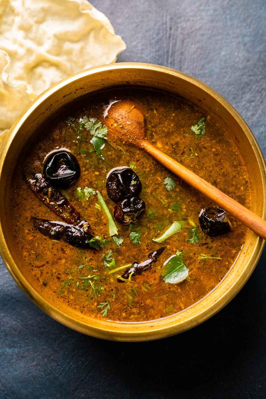 Picture of rasam in the brass pot that it was cooked in with a wooden ladle and some papad on the side