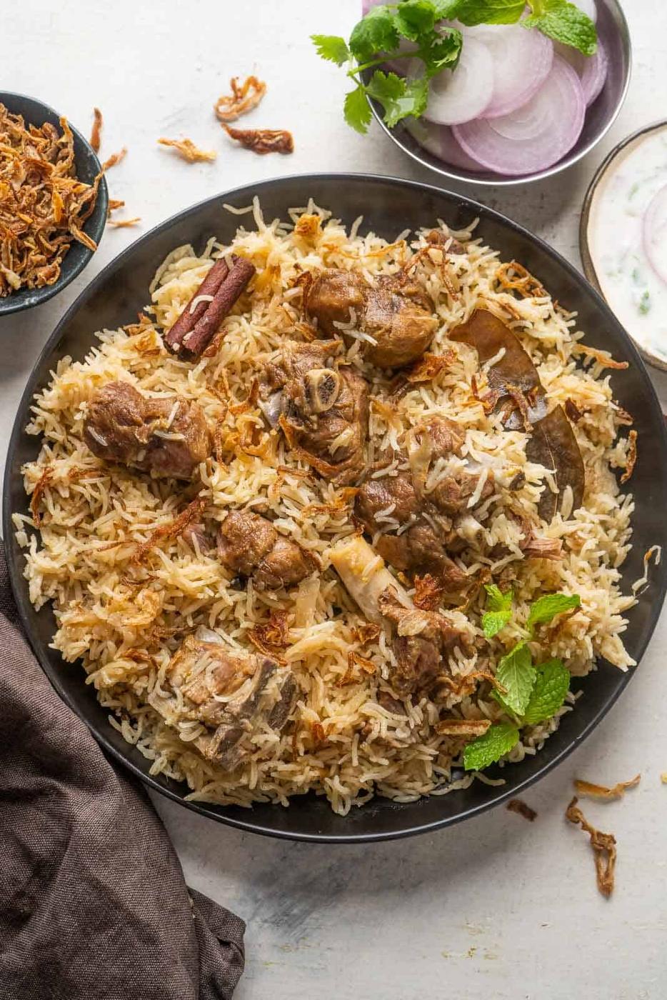 Mutton yakhni pulao served on a black plate with onions and raita on the side
