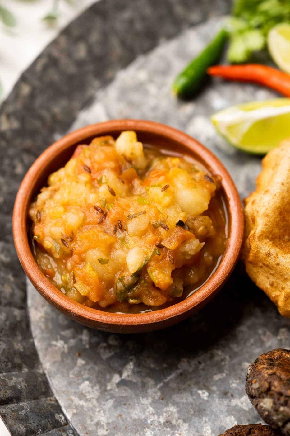 Picture of potato curry served in a brown terracotta bowl with puri on the side
