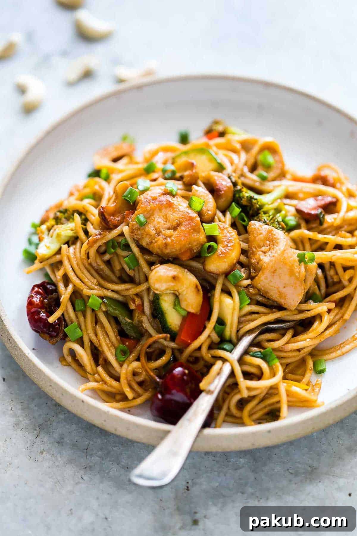 A close-up shot of the finished Chinese Cashew Chicken Noodles Stir-Fry served in a pristine white bowl, beautifully garnished with toasted sesame seeds and finely chopped green onions, ready to be enjoyed.