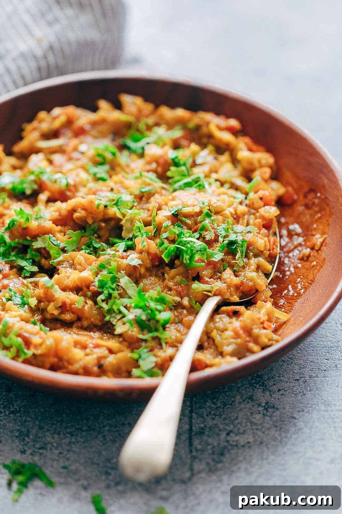 A close-up of a vibrant Baingan Bharta garnished liberally with fresh green coriander, served in a rustic red Indian dish. The texture and rich color of the dish are highly appealing, ready for consumption.