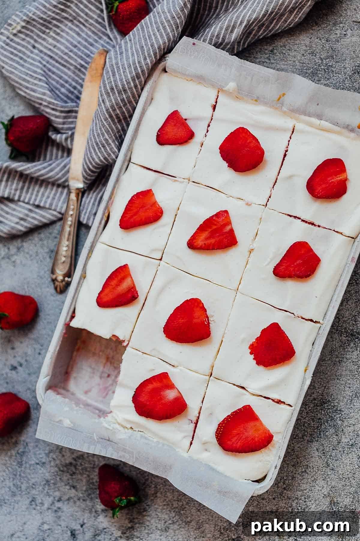 This from-scratch strawberry poke cake is incredibly soft, moist, and infused with jello, topped with whipped cream and fresh strawberries. An easy, elegant dessert perfect for any occasion. #dessert #cake #strawberry #easter #mothersday #pink A sheet pan filled with a freshly baked strawberry poke cake, partially sliced, with a serving knife resting next to it. Fresh whole strawberries are scattered around the pan, emphasizing the key ingredient.