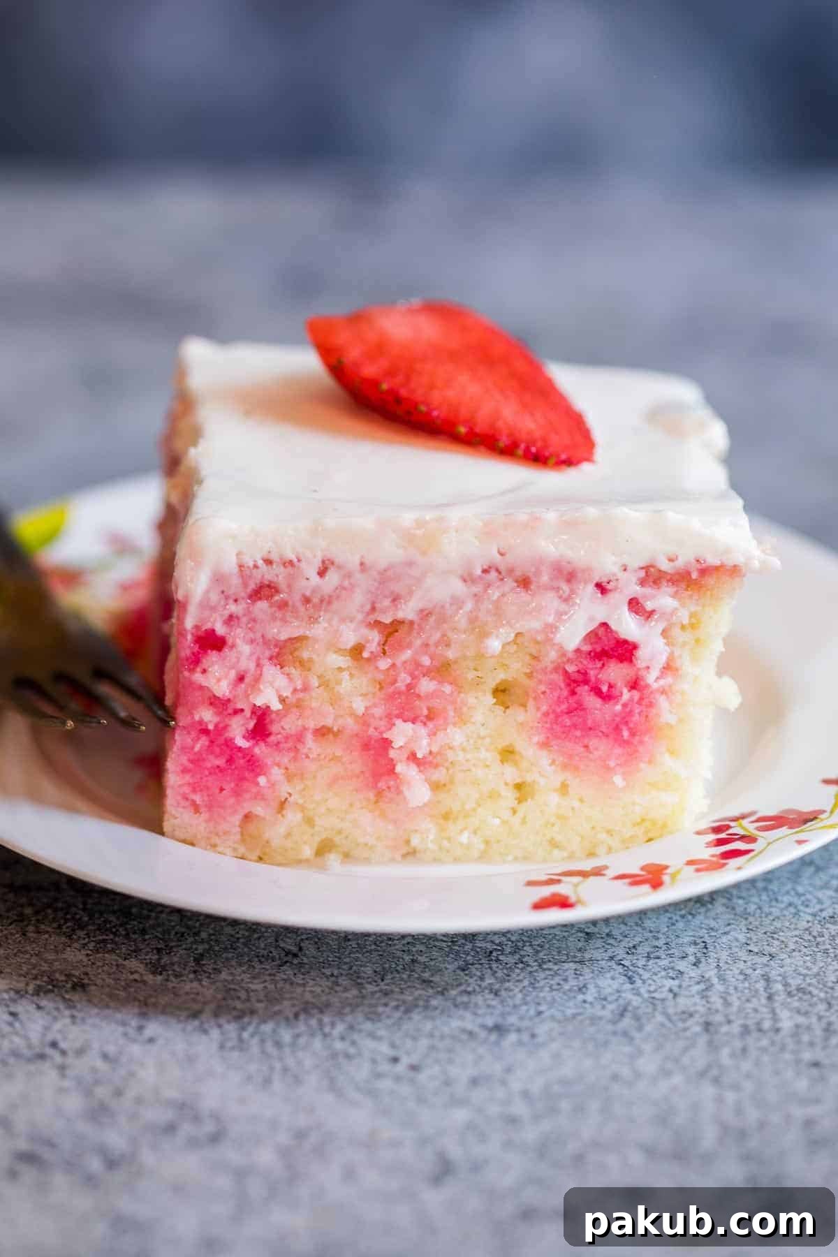 A delicious homemade strawberry poke cake, soft, moist, and infused with jello, topped with whipped cream and fresh strawberries. It's the perfect easy, elegant dessert for any occasion, delivering strawberry flavor in every bite. A close-up shot of a single slice of strawberry poke cake on a white plate, showcasing the vibrant pink jello streaks running through the moist cake layers, topped with creamy whipped cream and a fresh strawberry.