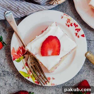A slice of homemade strawberry poke cake served on a white plate with a fork and fresh strawberries.