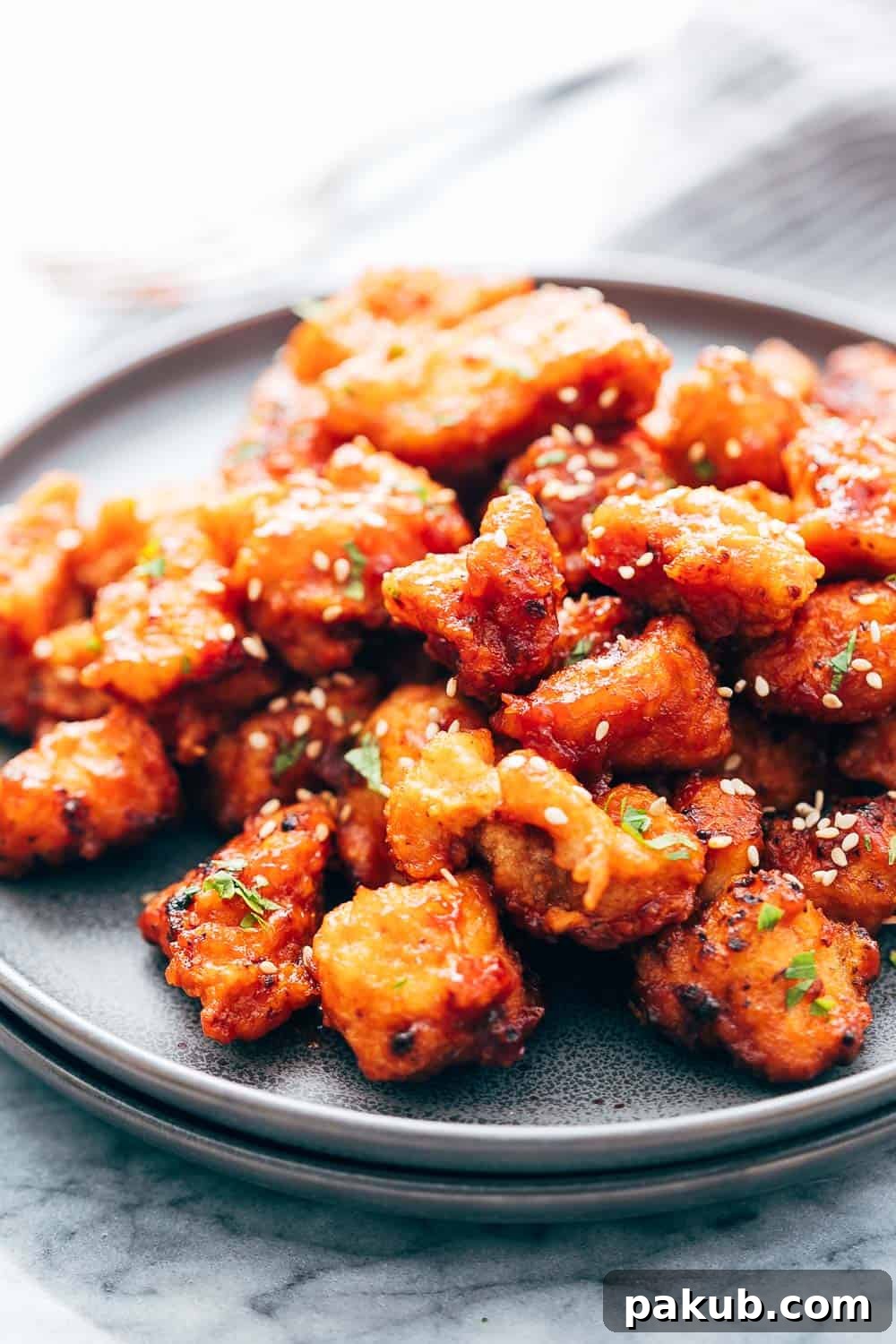 KOREAN POPCORN CHICKEN is sticky, spicy, tangy and crunchy. Inspired by the hyper-famous Korean street snack, Dakgangjeong, boneless chicken is deep fried and tossed in a sweet, sour and spicy sauce that makes every bite super addictive. #chicken #appetizer #party #snack #recipe Closeup of Korean Popcorn Chicken on a grey ceramic plate topped with sesame seeds and chopped parsley