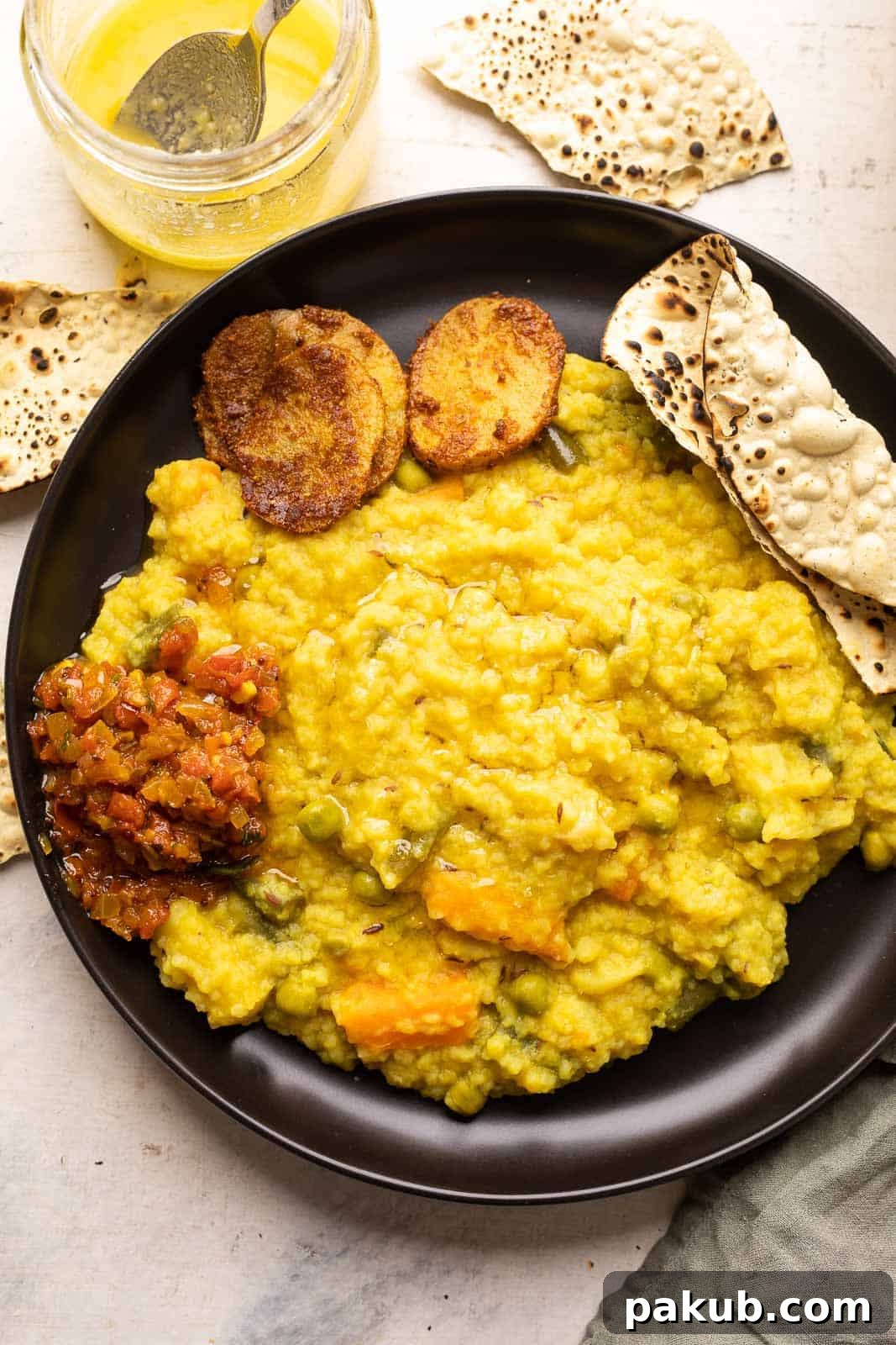 Irresistibly Delicious Khichdi 2 Khichdi served on a black plate with papad, tomato chutney and aloo fry