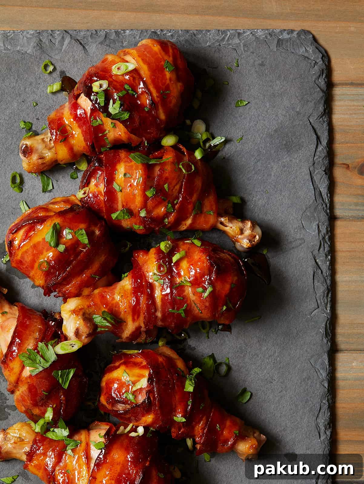 15 Bacon Creations to Make Every Meal a Party 11 Sticky and savory Maple Bacon Chicken Drumsticks artfully arranged on a sleek slate serving board, showcasing their beautiful glaze and crispy bacon wrapping.
