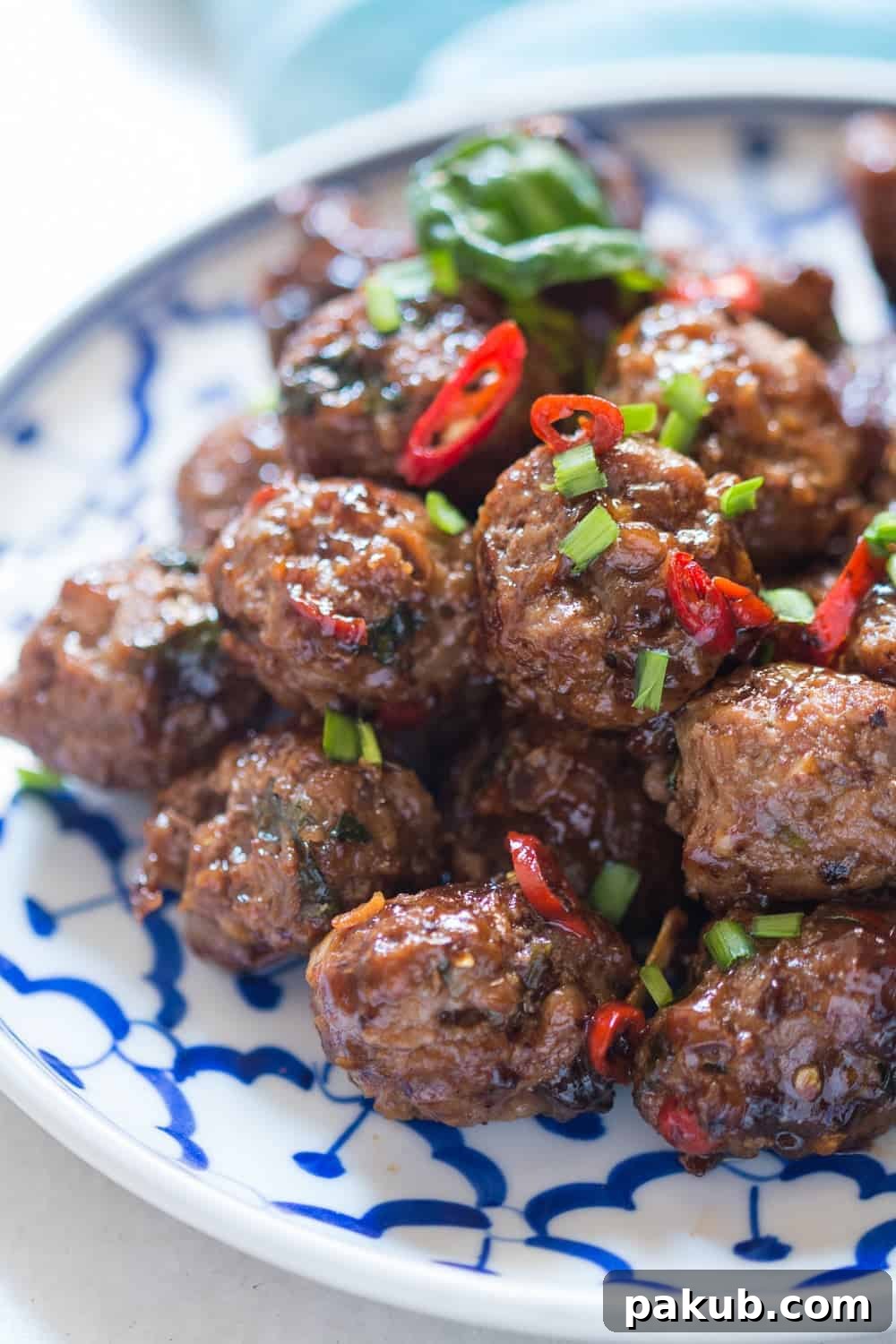close up shot of sticky meatballs garnished with spring onions and thai red chillies served in a white and blue plate