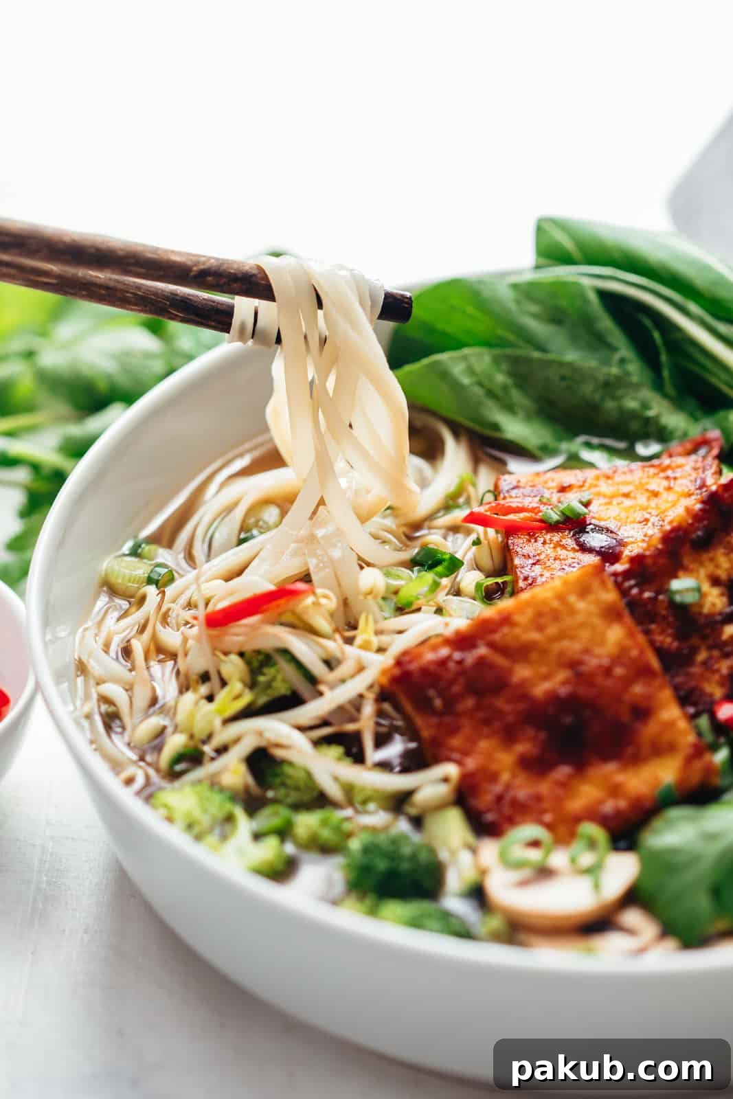 Plant-Powered Pho Bowl 4 Chopsticks lifting flat rice noodles from a steaming bowl of vegetarian pho, surrounded by colorful garnishes.