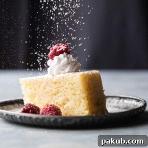 A slice of eggless vanilla cake with whipped cream, raspberries and a sprinkling of icing sugar