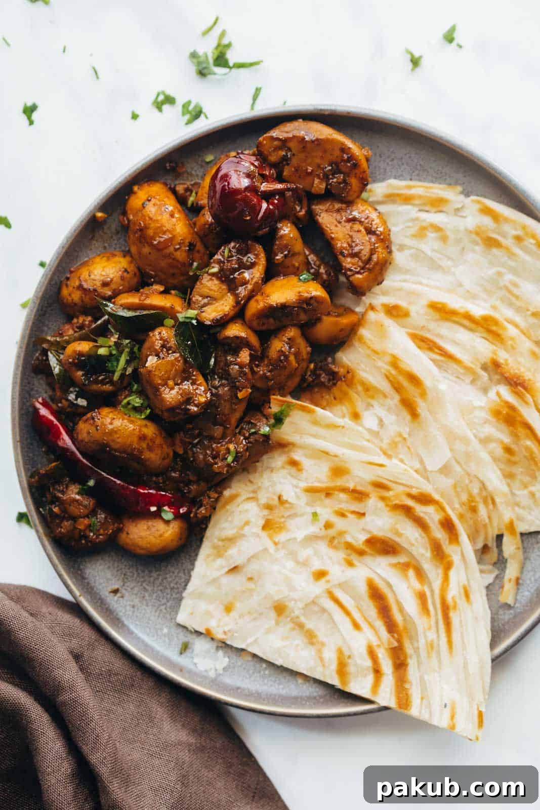 Peppery South Indian Mushroom Fry 2 Delicious South Indian Mushroom Pepper Fry served on a grey plate with traditional Malabar Parathas, showcasing its rich texture and inviting aroma.