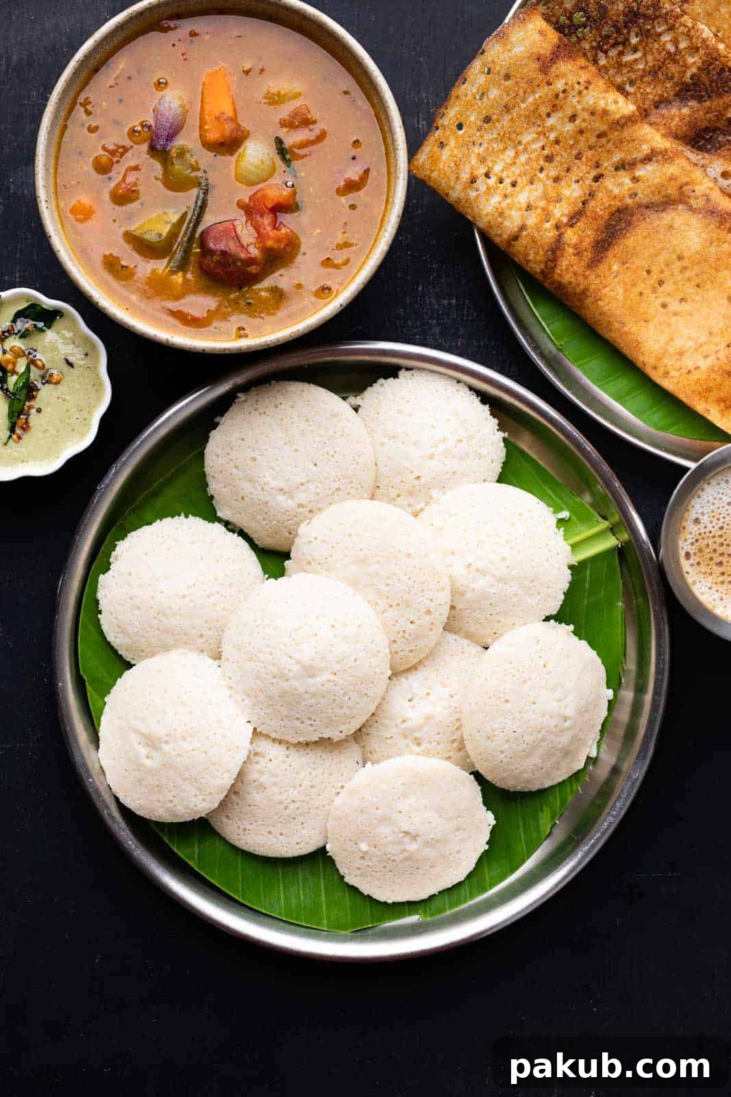 A stack of soft, fluffy idlis on a banana leaf-lined plate, accompanied by crispy dosas, fresh coconut chutney, and aromatic sambar, showcasing a complete South Indian breakfast.