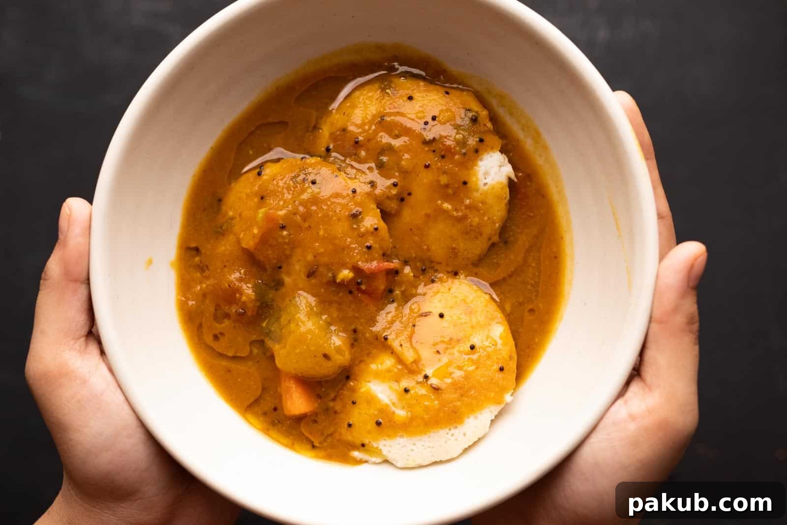 Sambar poured over steaming hot idlis in a bowl, with hands holding the bowl as if serving someone.
