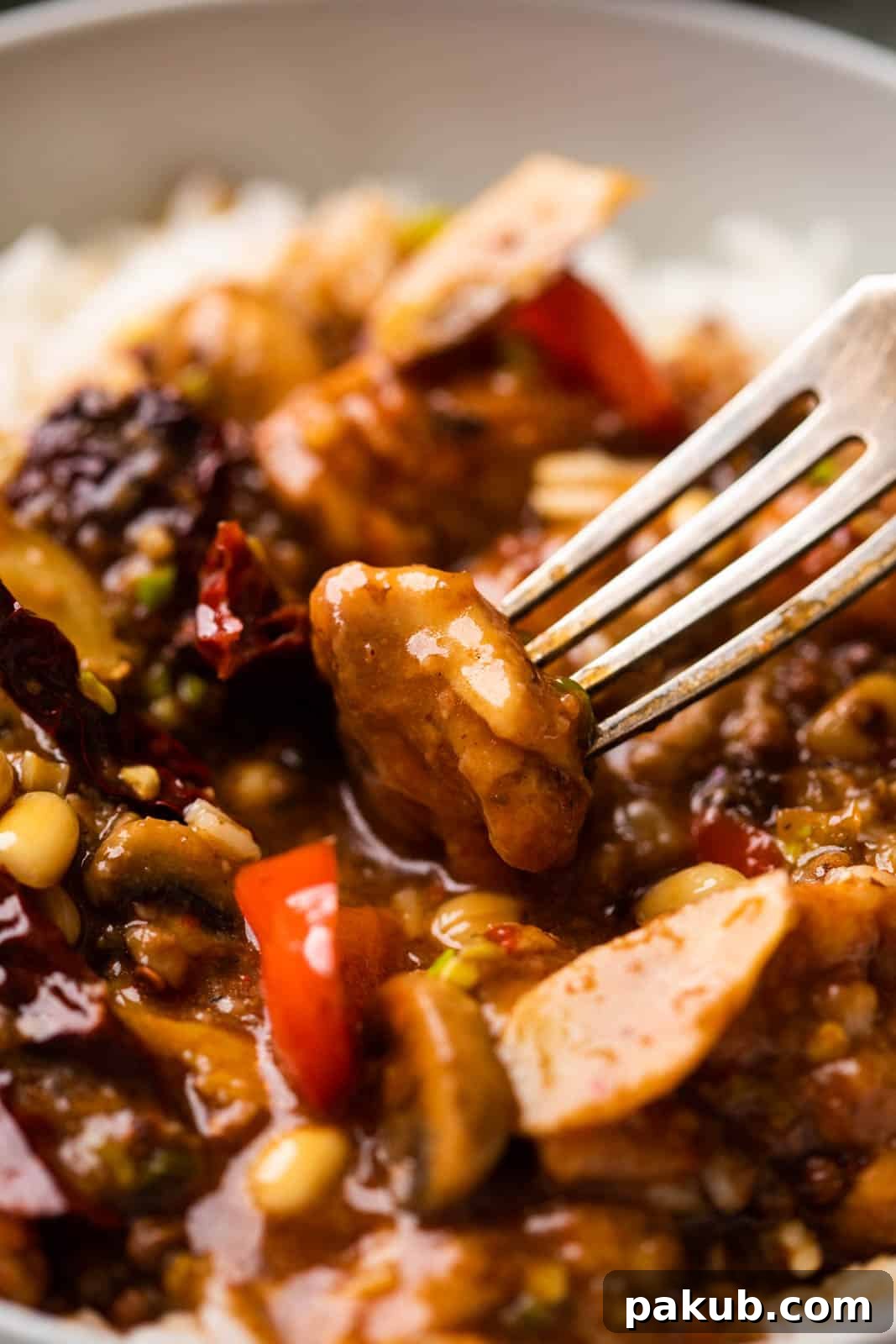 A close-up shot of a single piece of Kung Pao chicken, glistening with sauce, on a fork.