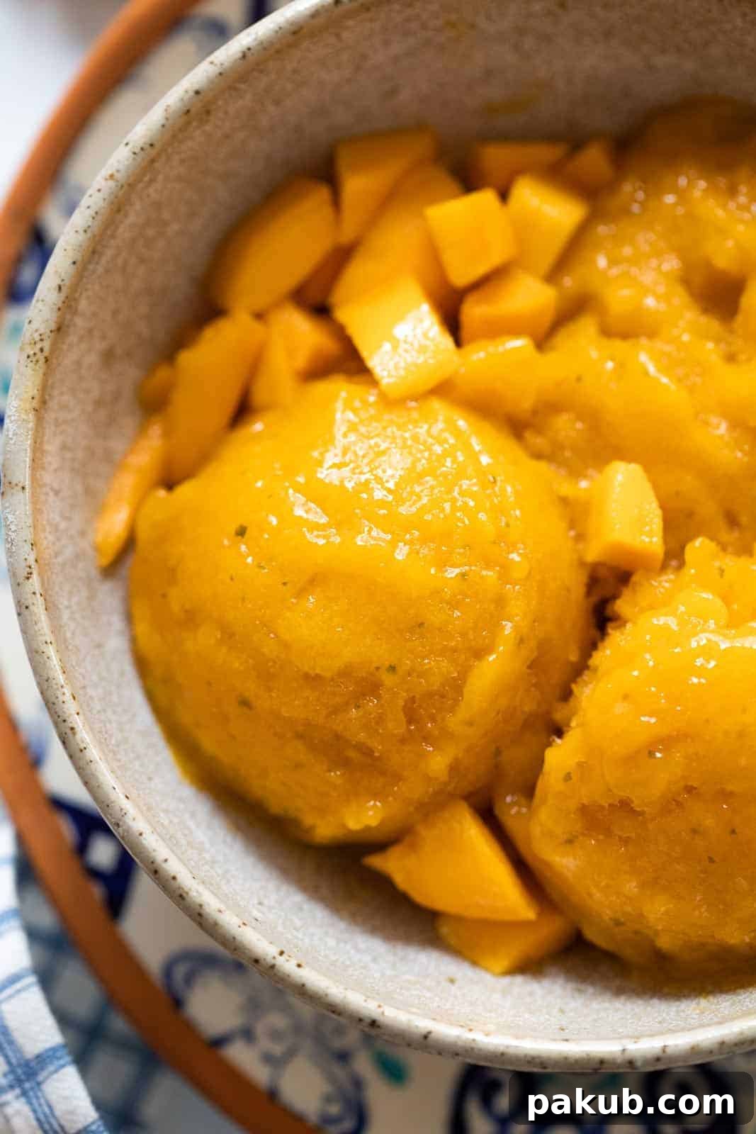 Whip Up Homemade Mango Sorbet Without a Machine 2 Scoops of homemade Alphonso mango sorbet served in a rustic brown ceramic bowl, garnished with fresh diced mangoes, capturing the vibrant golden color and smooth texture of the dessert.