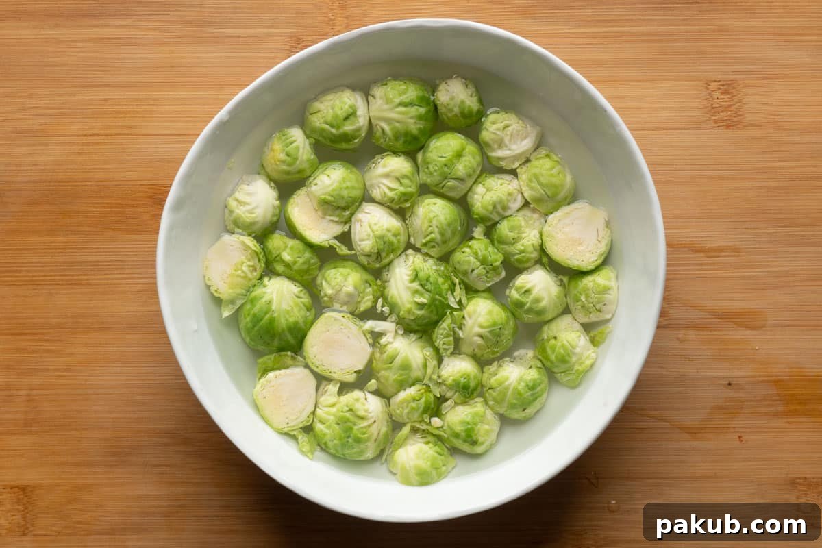 A bowl filled with trimmed and halved Brussels sprouts, submerged in clean cold water, illustrating the crucial soaking step designed to ensure optimal internal tenderness during air frying.
