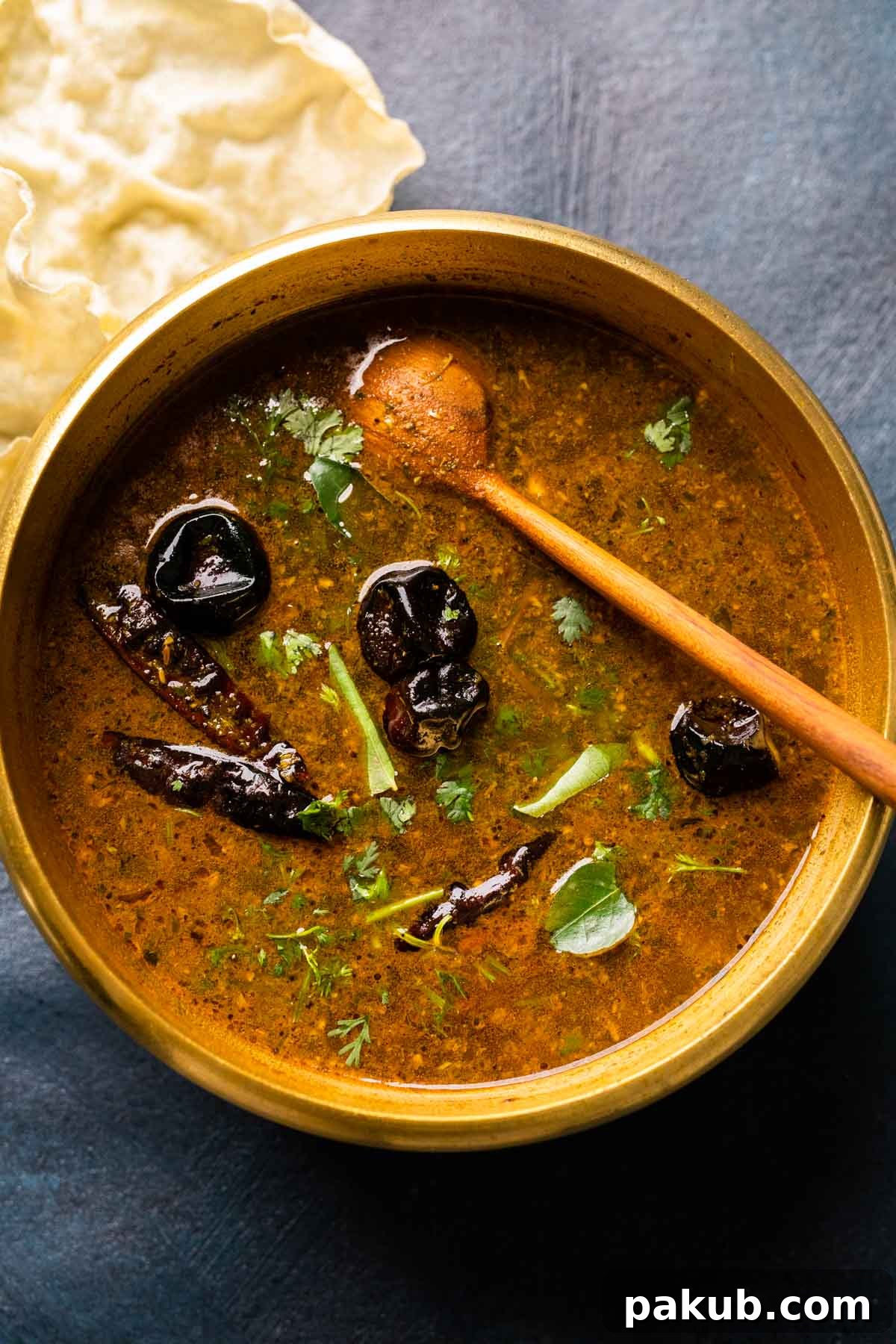 Picture of rasam in the brass pot that it was cooked in with a wooden ladle and some papad on the side