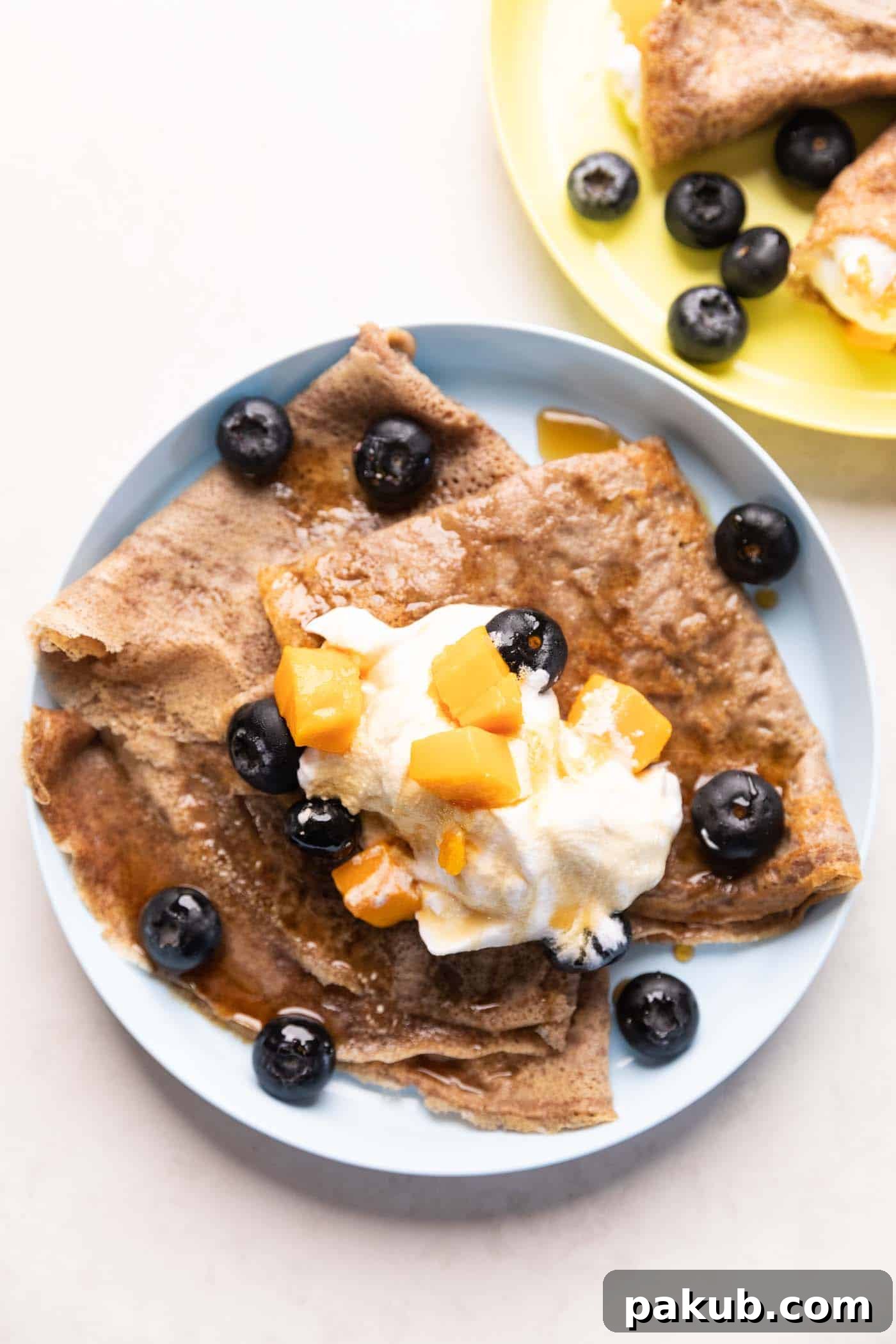 Delicious Ragi Crepes served on a plate, beautifully topped with fresh whipped cream, juicy mango slices, and vibrant blueberries, ready to be enjoyed.