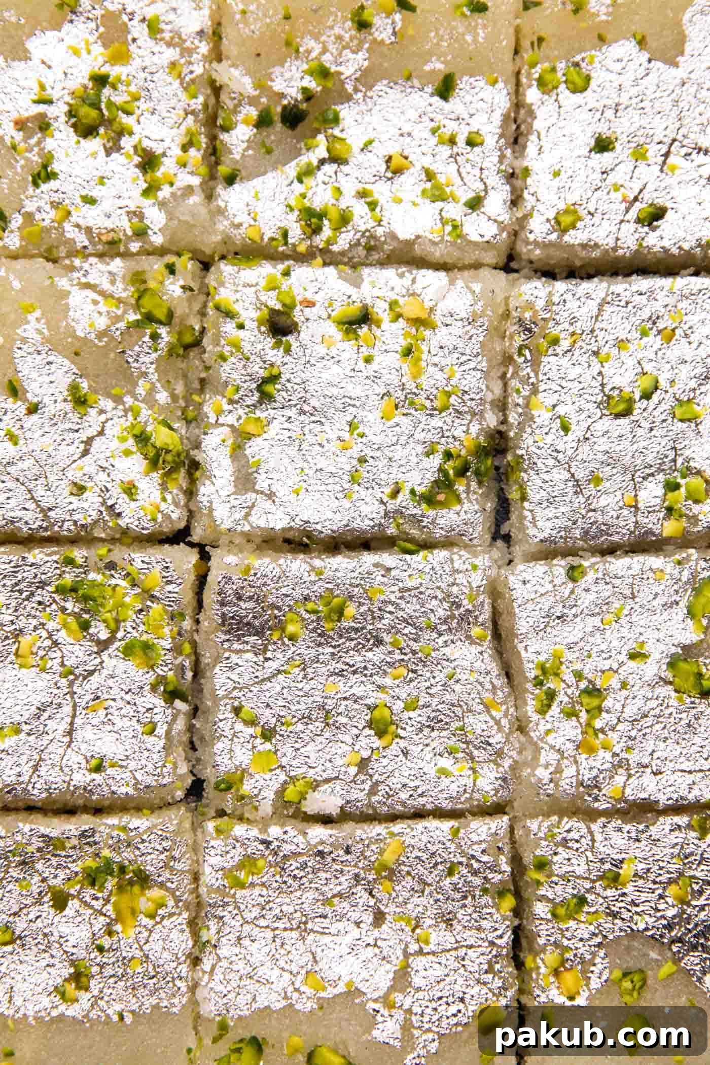Sweet Coconut Fudge 6 A close-up view of perfectly cut Coconut Barfi squares, glistening with edible silver leaf and speckled with chopped pistachios, signaling they are ready to be savored.