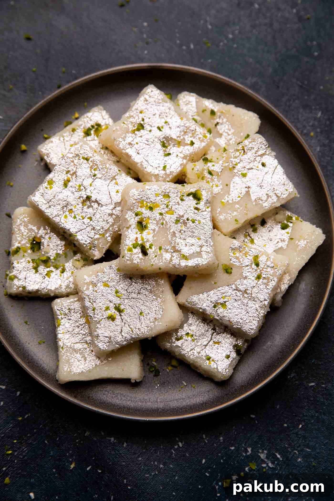Sweet Coconut Fudge 7 Coconut Barfi squares beautifully presented on a sleek grey plate, adorned with a final sprinkle of pistachios, eagerly awaiting to be enjoyed as a festive dessert.