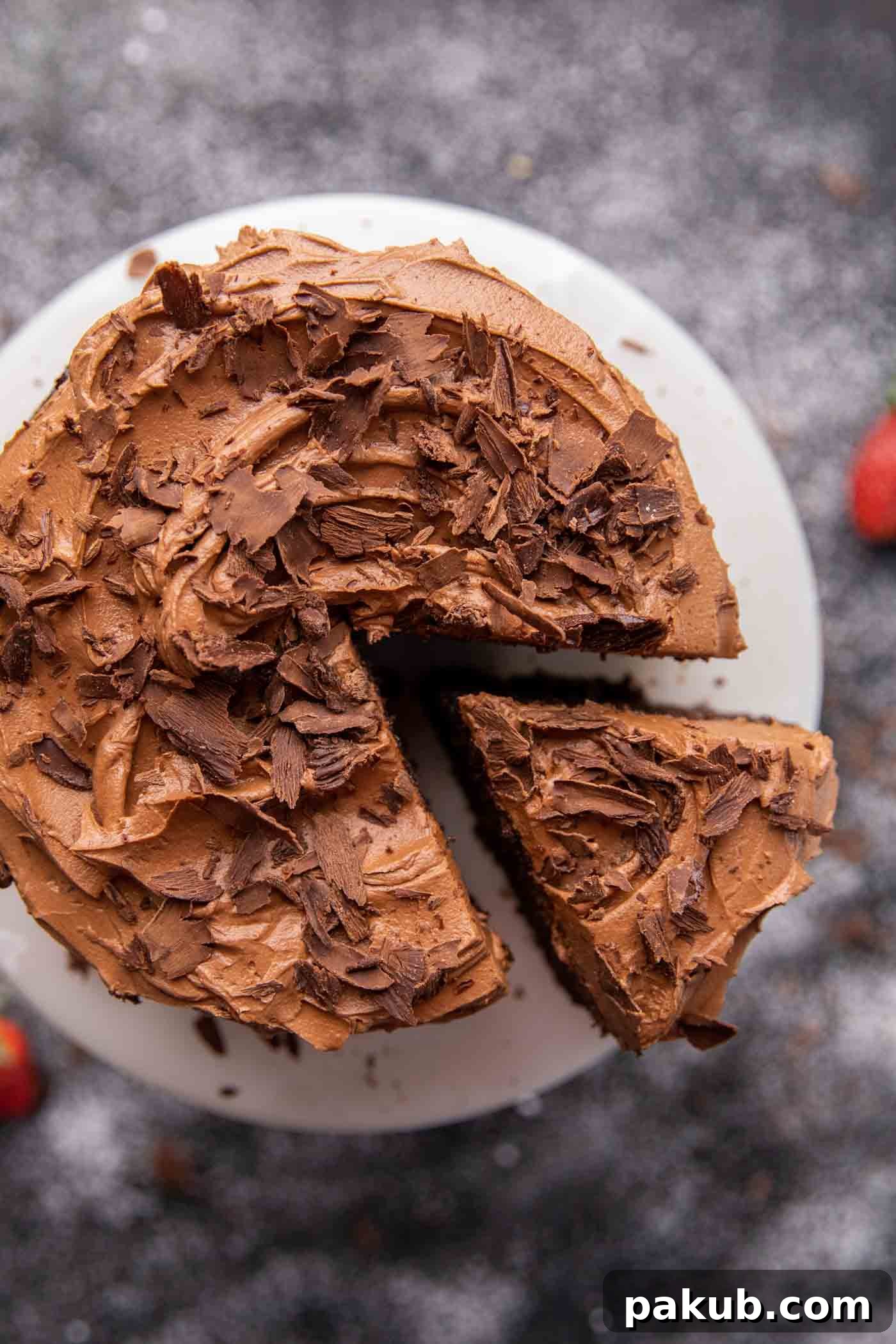 A lush, moist chocolate cake on a white cake stand, generously topped with smooth chocolate buttercream frosting and delicate chocolate flakes.