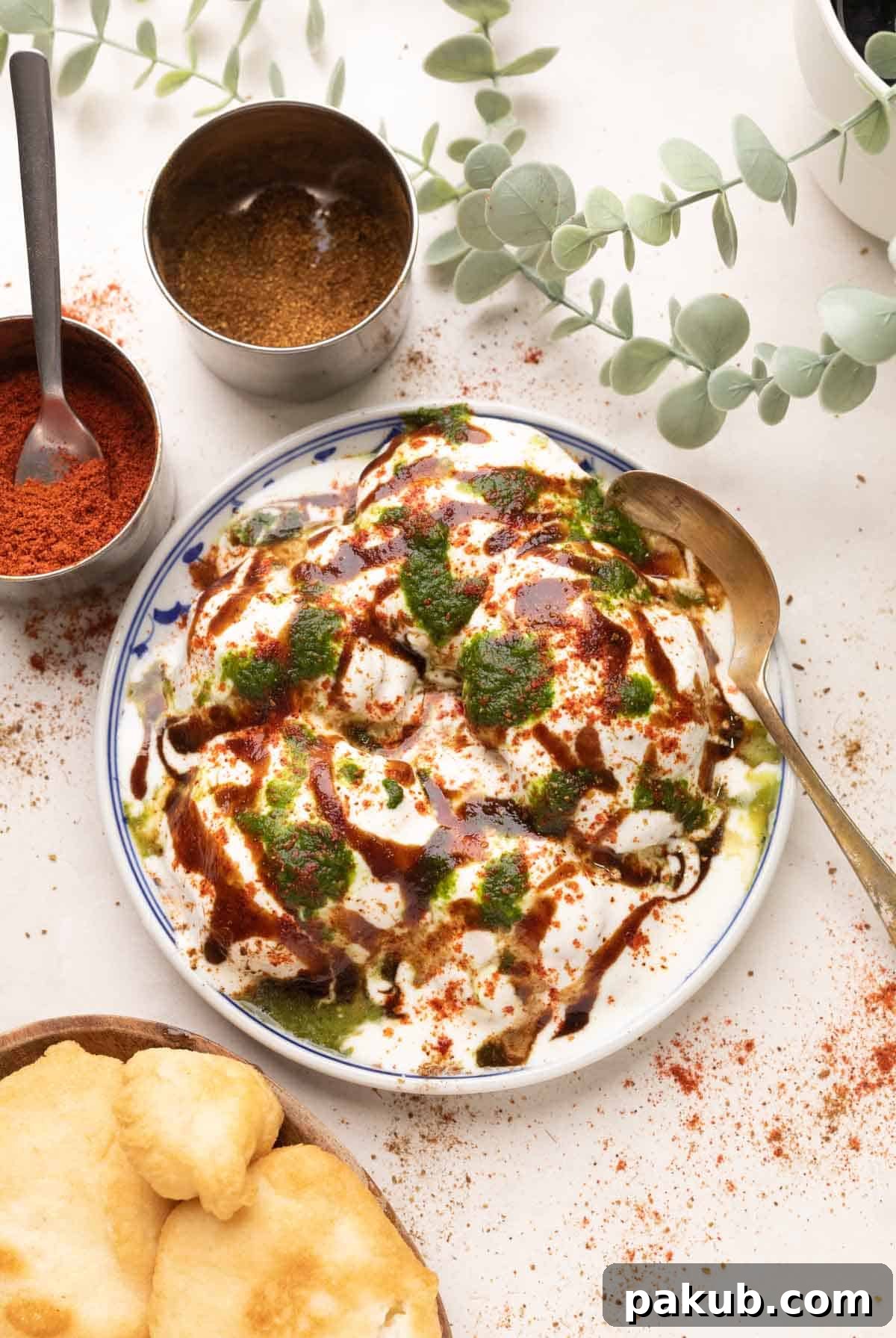 Cloud-Soft Lentil Fritters in Spiced Yogurt 9 A beautifully arranged plate of Dahi Vada, generously drizzled with tamarind chutney, coriander mint chutney, and a sprinkle of spices, with additional vadas and ingredients in the background.
