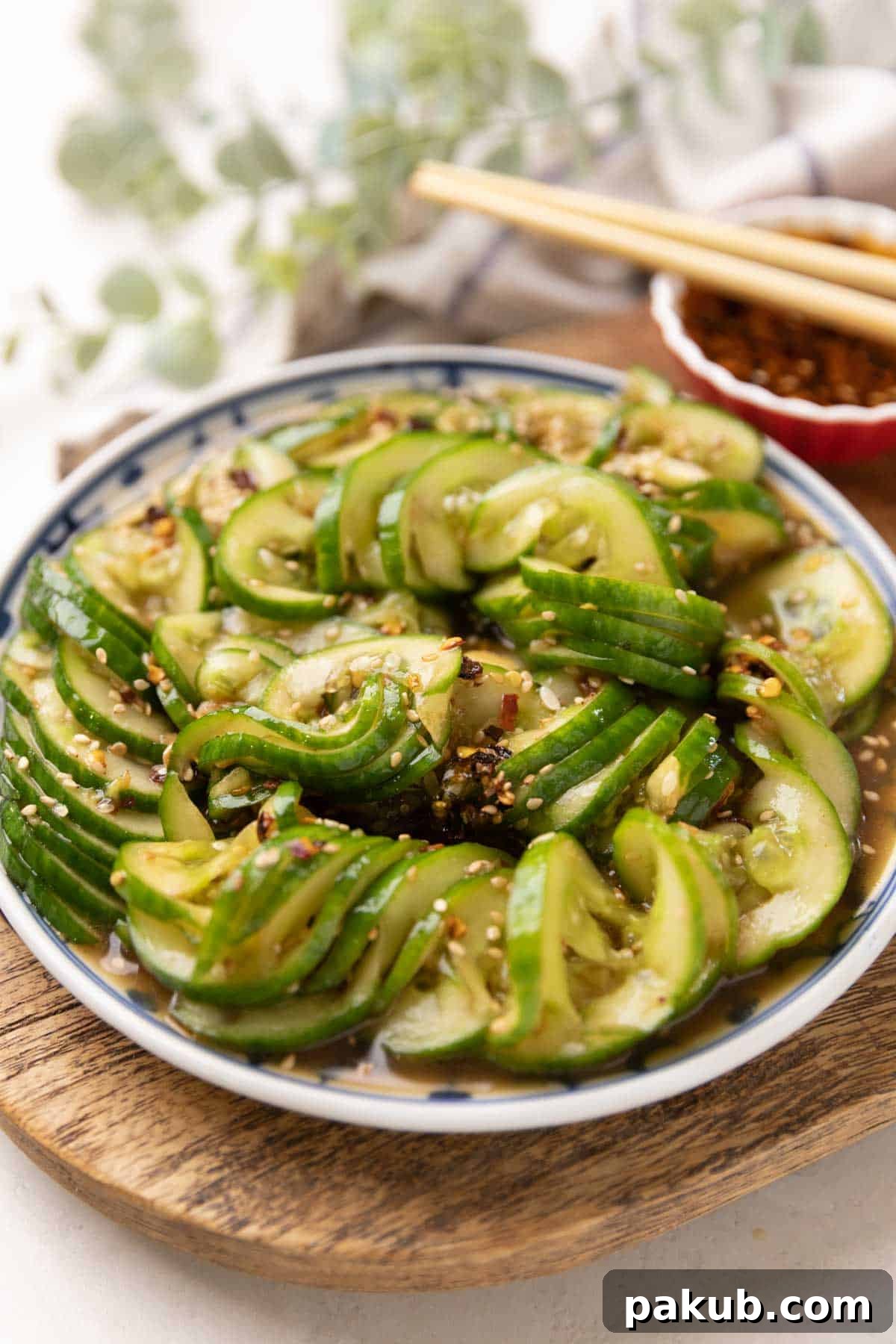 Sesame Garlic Cucumber Salad 4 An elegantly presented Asian cucumber salad in a blue and white plate on a wooden board, accompanied by a small red bowl of dressing and traditional chopsticks.