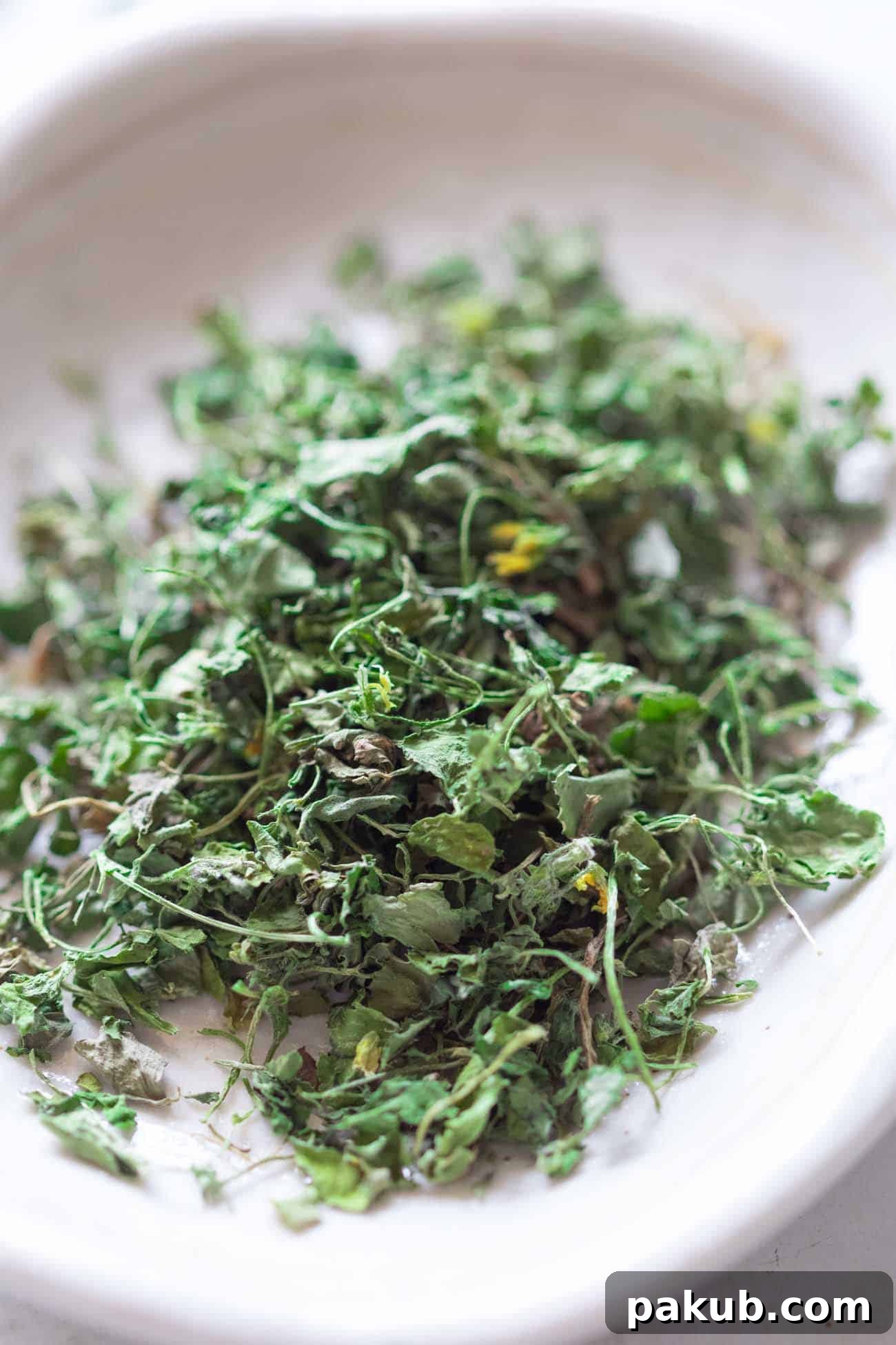 Kasuri Methi Your Complete Guide to Dried Fenugreek Leaves and Their Uses 3 A closer, detailed shot of sun-dried Kasuri Methi leaves, crumbled on a white plate, highlighting their texture and readiness for use as an aromatic garnish or ingredient.