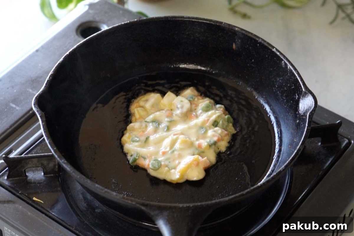 A savory vegetable pancake sizzling in a hot skillet, with a perfectly golden-brown and crispy bottom, while the vibrant mixed vegetables are visible within the batter, signifying it's halfway cooked.