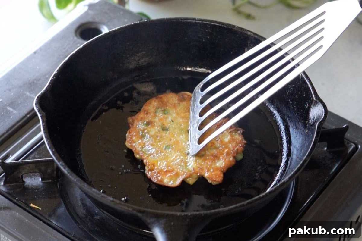A perfectly cooked golden-brown vegetable pancake being gently pressed with a spatula in a hot skillet, ensuring optimal crispiness and even cooking on both sides.