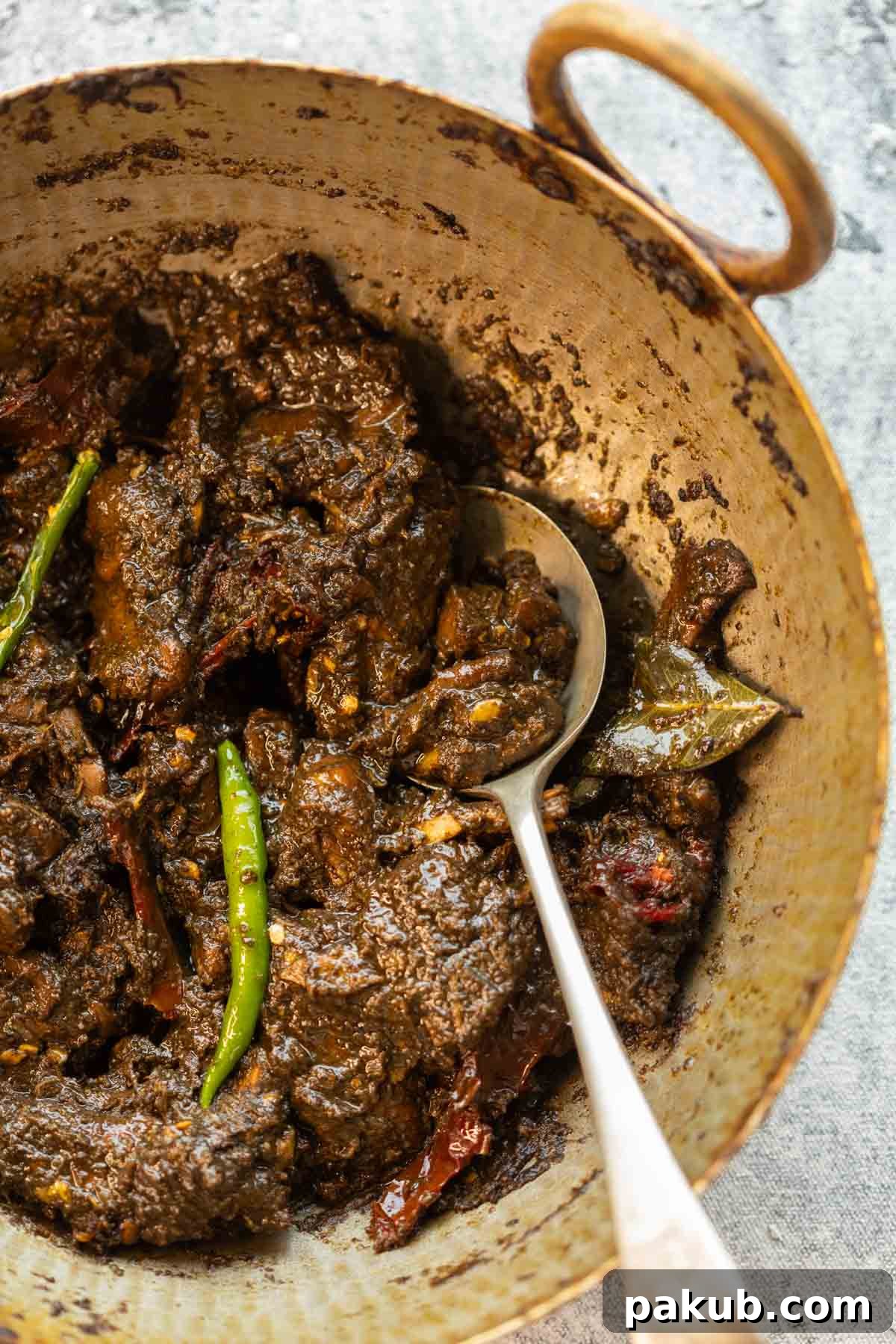 Black sesame chicken curry served in a traditional brass kadhai with a spoon, inviting you to taste this unique Northeast Indian dish.