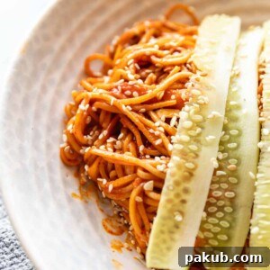 A refreshing Bibim Guksu (Spicy Korean Noodles Salad) expertly garnished with crisp cucumber, served elegantly in a pristine white bowl, promising a delightful culinary experience.