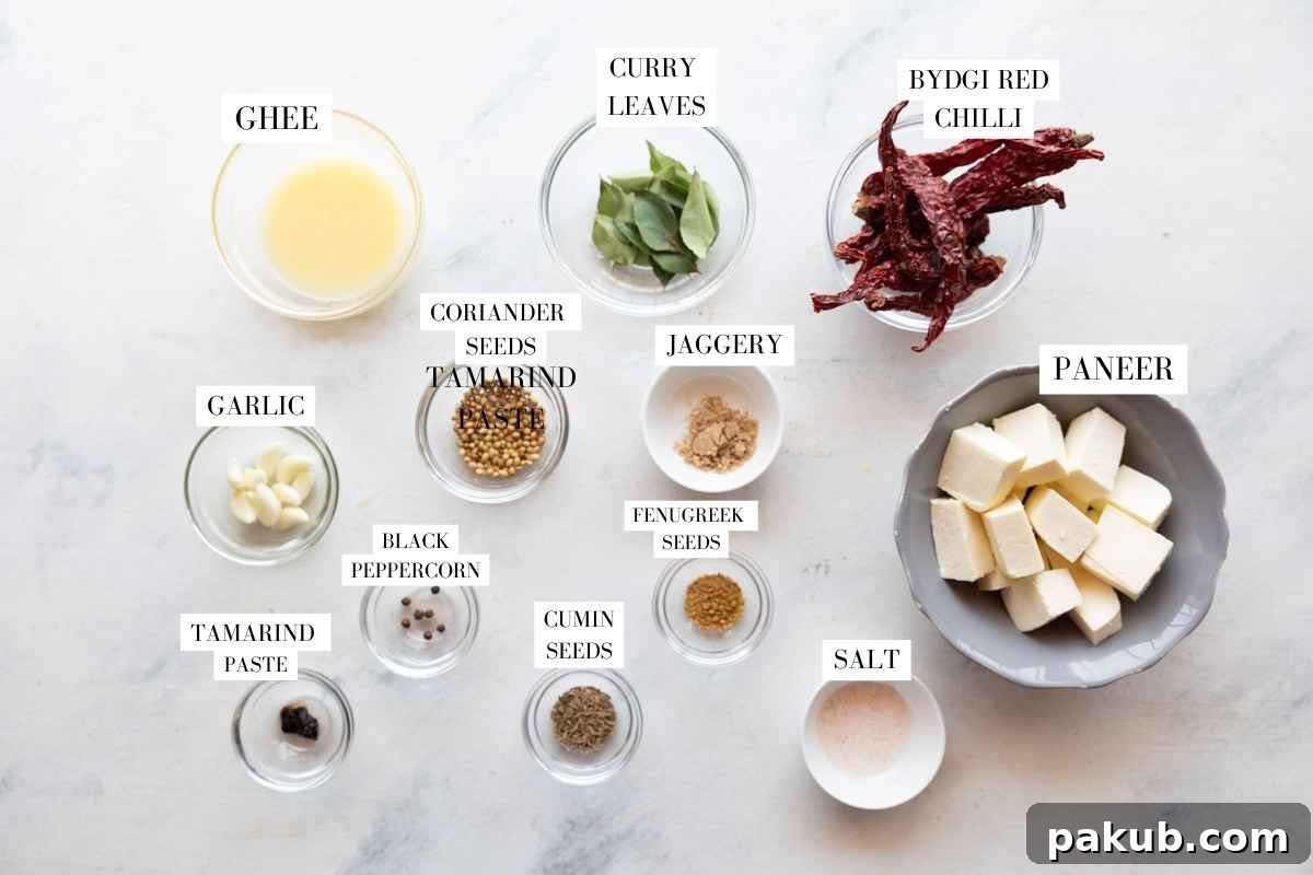 A beautifully arranged spread of fresh ingredients for Paneer Ghee Roast on a pristine white surface, including golden paneer cubes, rich ghee, whole spices like coriander and cumin, bright red Byadgi chillies, and garlic cloves.