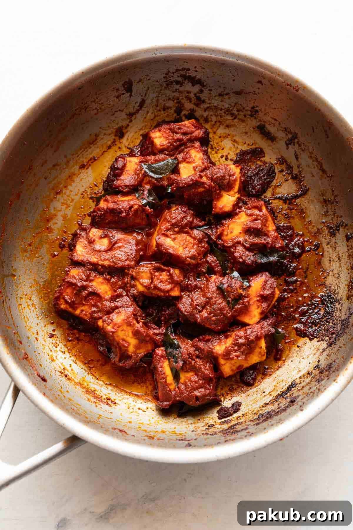 A close-up view of the Paneer Ghee Roast simmering in a stainless steel kadhai, showcasing the vibrant reddish-maroon color and the rich, glossy texture of the curry as it reaches perfection.