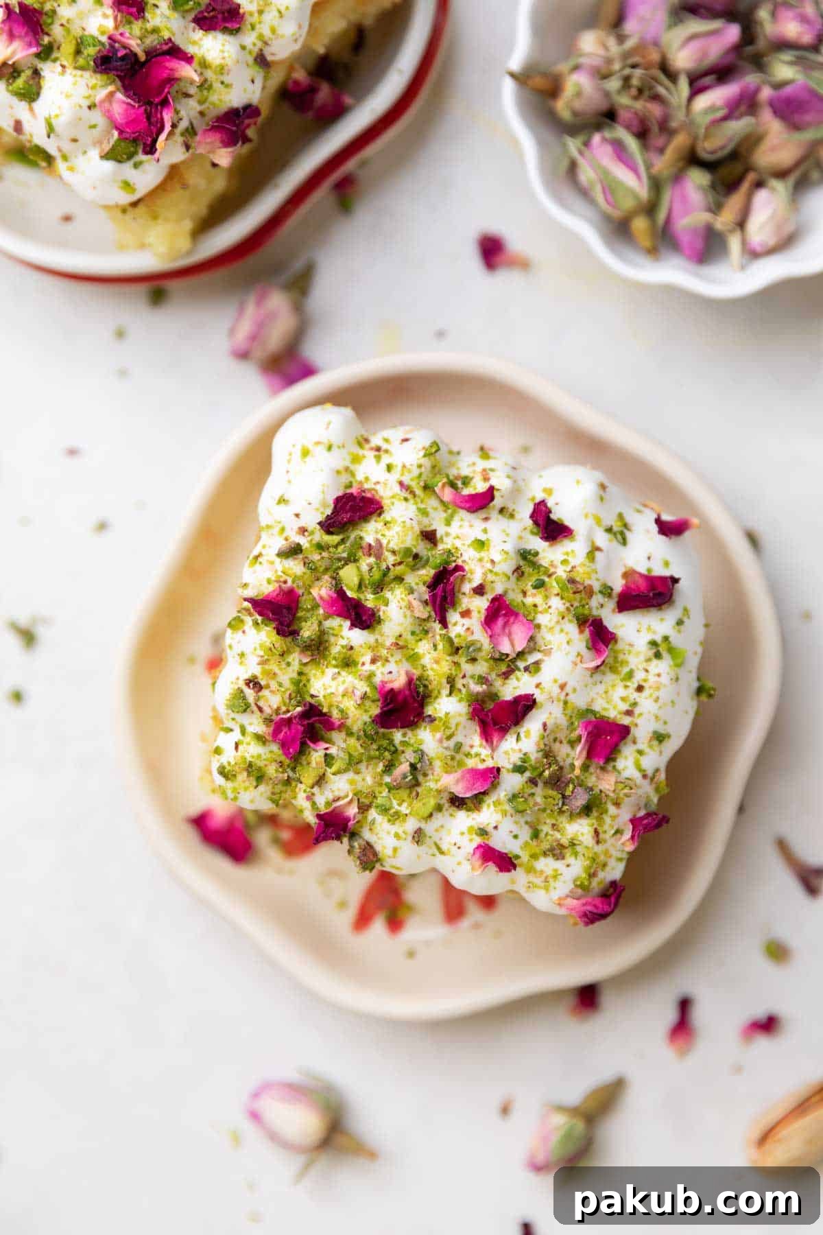 A beautifully garnished slice of rasmalai cake on a white plate, adorned with whipped cream, crushed pistachios, and delicate rose petals.