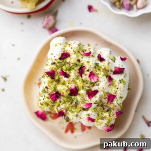 A slice of rasmalai cake garnished with cream, crushed nuts and rose petals on a white plate