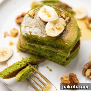 Stacked spinach banana pancakes topped with banana slices, walnuts, and honey, served with a fork.