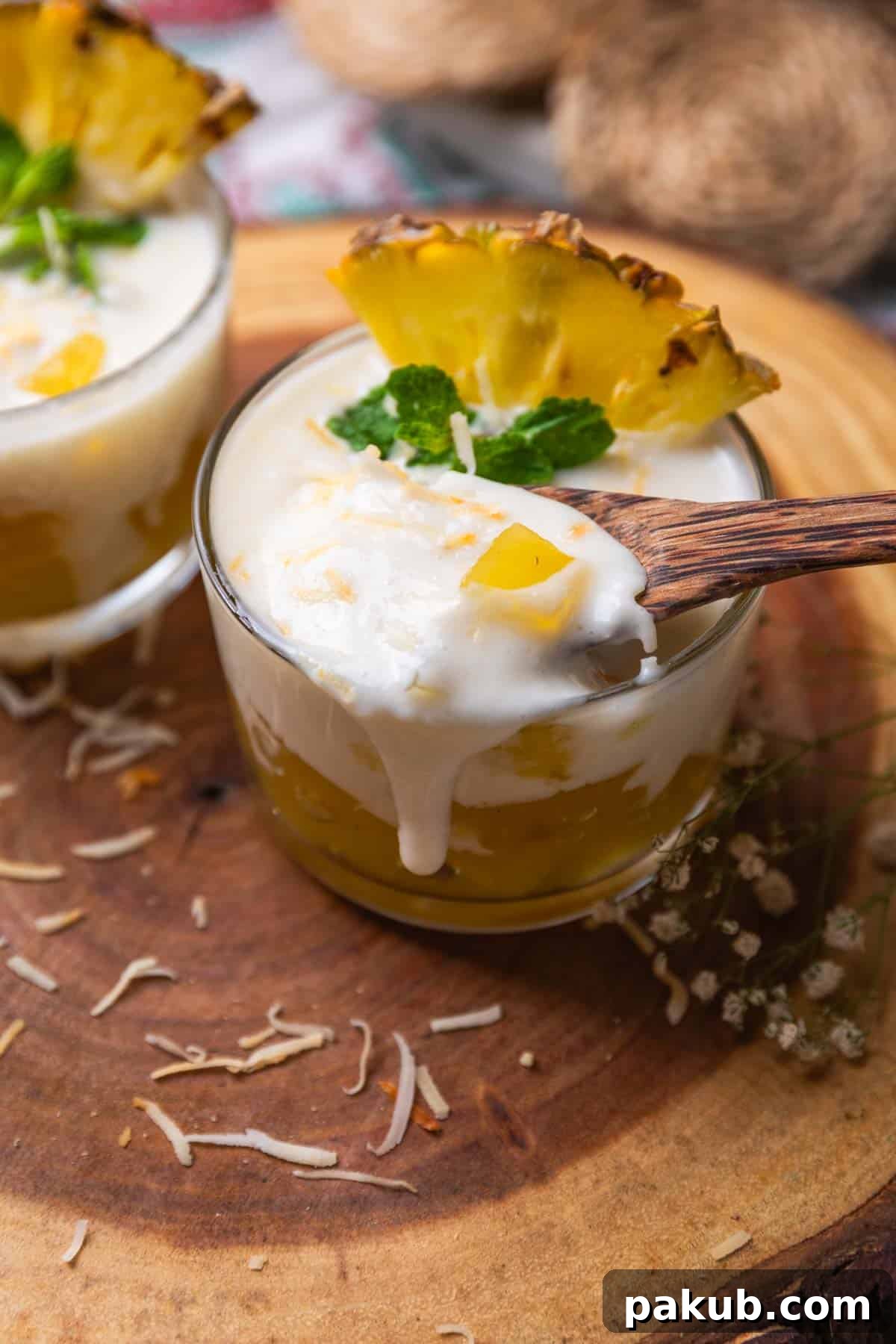 coconut pudding with pineapple jelly garnished with toasted coconut flakes, pineapple slice and mint leaves and served in shot glasses and a wooden spoon
