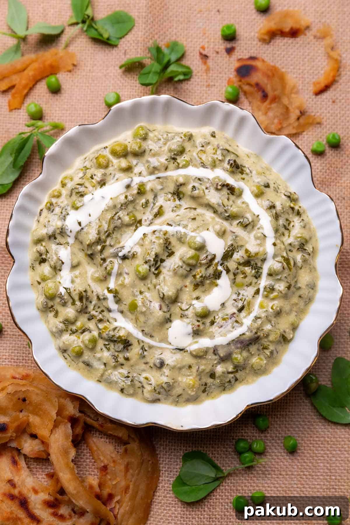 A beautifully presented bowl of creamy Methi Matar Malai, garnished with fresh fenugreek leaves and bright green peas, served alongside a flaky lachha paratha on a rustic wooden surface.