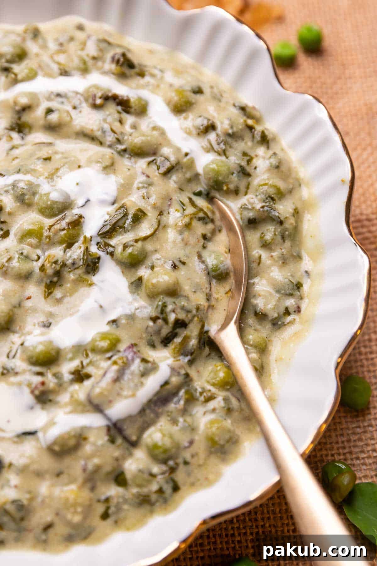 A close-up shot of a serving spoon lifting a generous portion of creamy Methi Matar Malai from a white bowl, clearly showing the rich gravy, tender green peas, and finely chopped fenugreek leaves.
