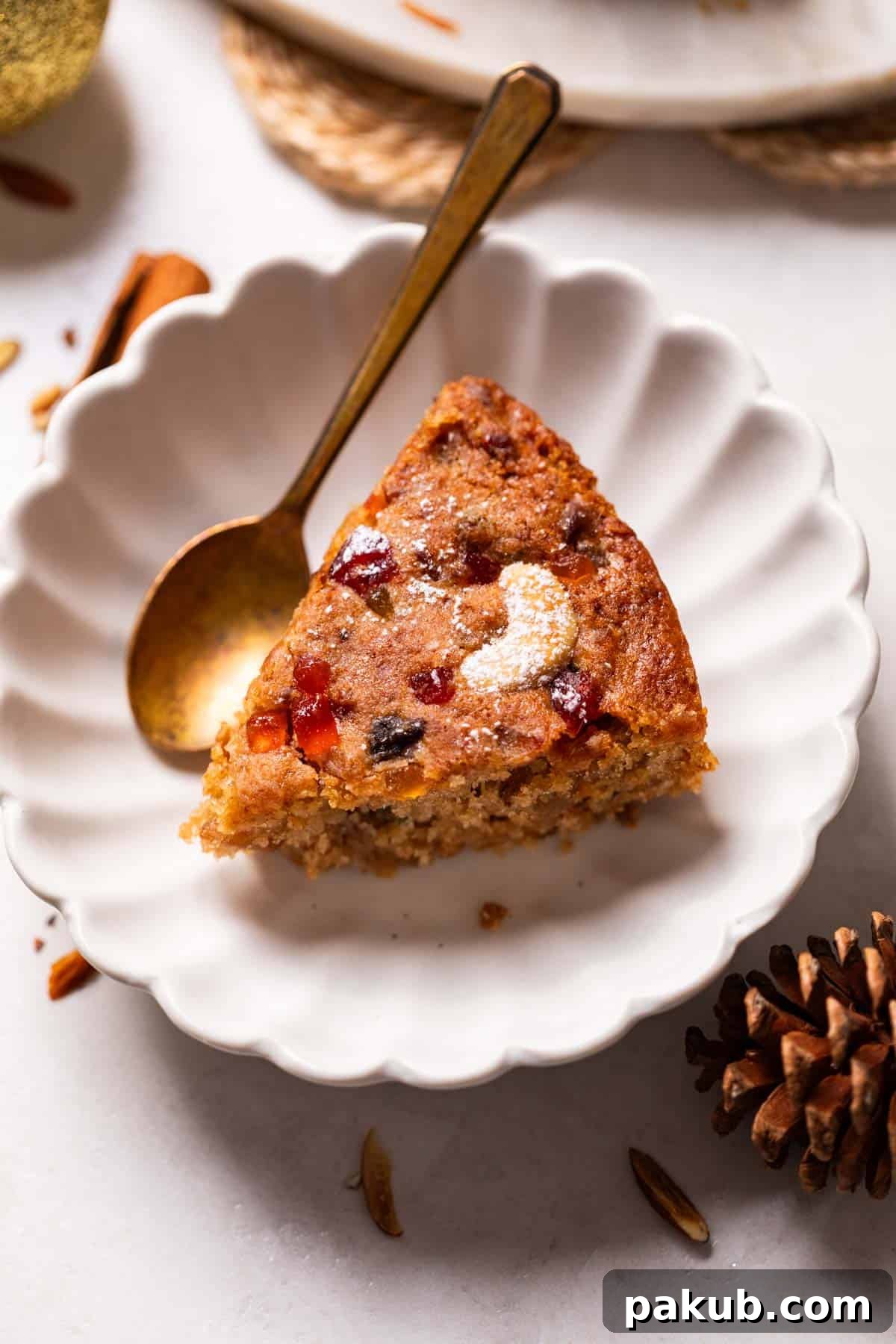 A perfectly baked slice of eggless plum cake, showing its moist crumb and abundance of fruits and nuts, served on a white dish with a spoon.