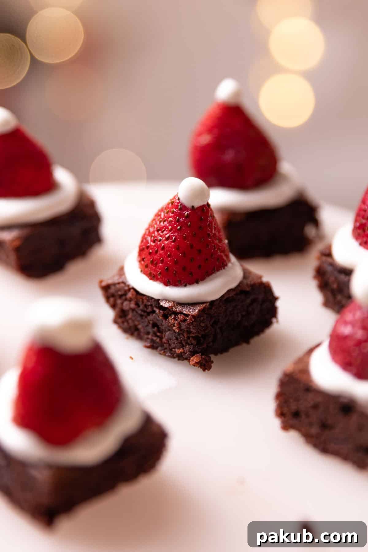 Close-up of festive brownie bites arranged on a pristine white platter, each adorned with a swirl of white frosting, a fresh strawberry cap, and a hint of mint, resembling mini Santa hats, perfect for holiday gatherings.