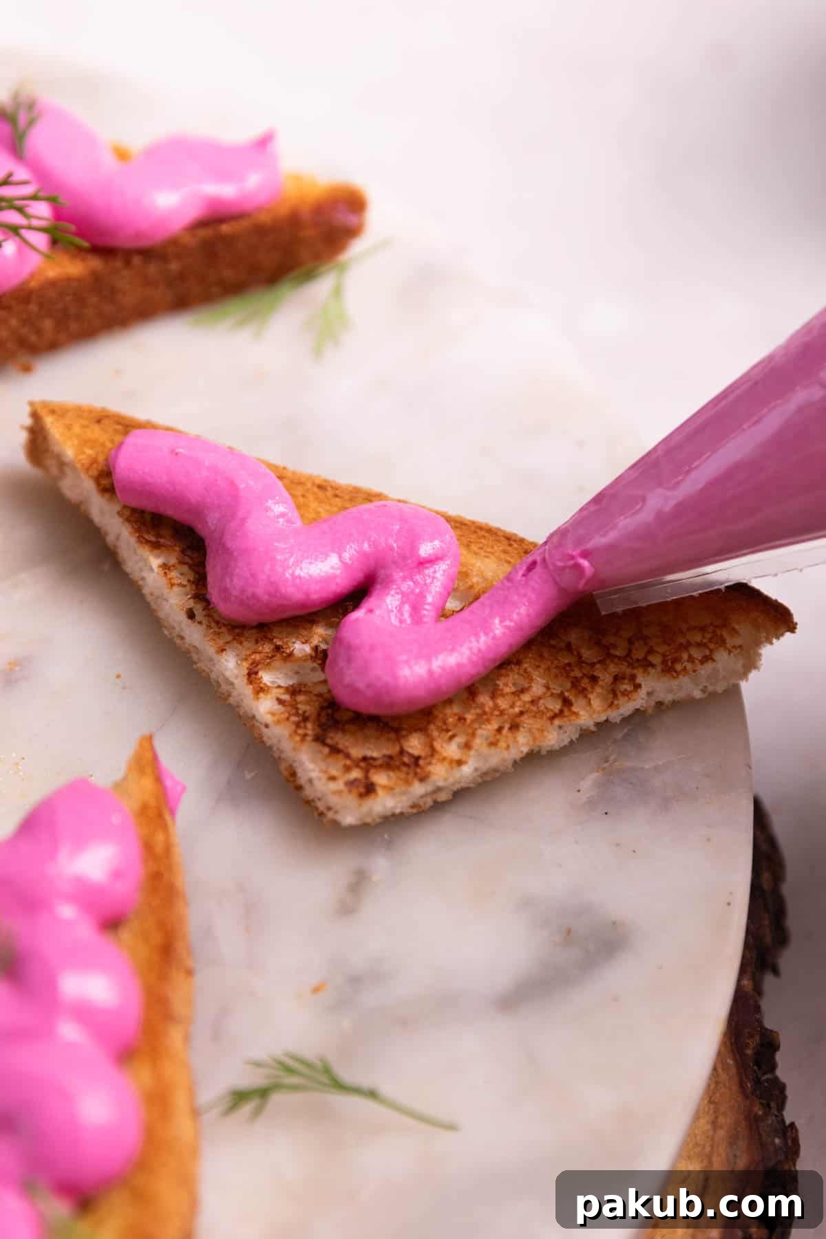 A close-up image illustrating the delicate act of piping creamy paneer beetroot mixture onto crisp, golden toast triangles, highlighting the enticing contrast between soft and crunchy textures.