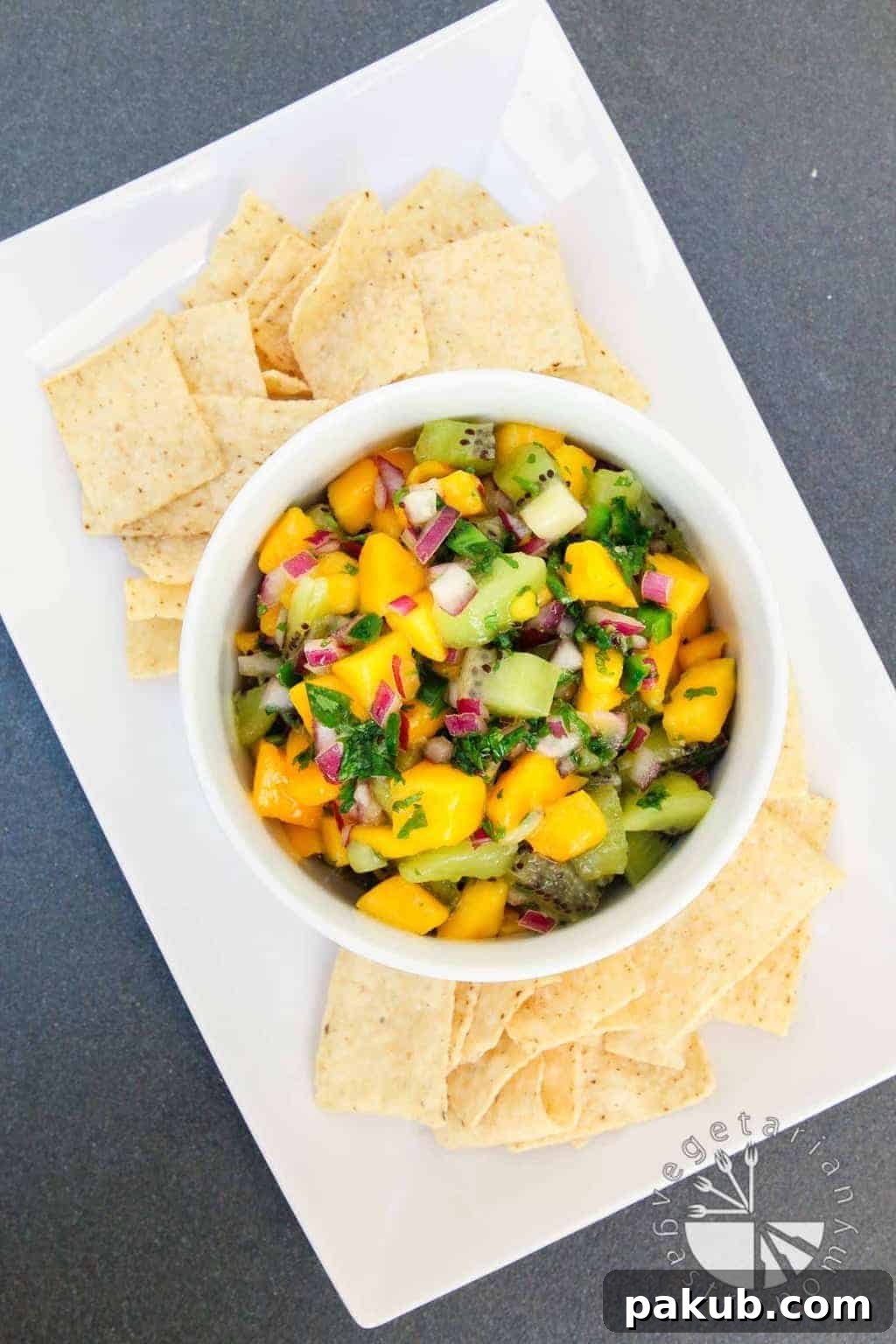 A collection of 13 insanely delicious savoury mango recipes that are sure to blow your mind if you've always had mangoes in sweet dishes. These recipes combine the juicy sweetness of mangoes with savoury ingredients to create a dish that's truly stellar and unforgettable! #spring #mango #recipes #easter #summer #fruit A brightly colored Kiwi Mango Salsa artfully arranged on a pristine white plate, accompanied by crisp tortilla chips, ready for a refreshing snack.