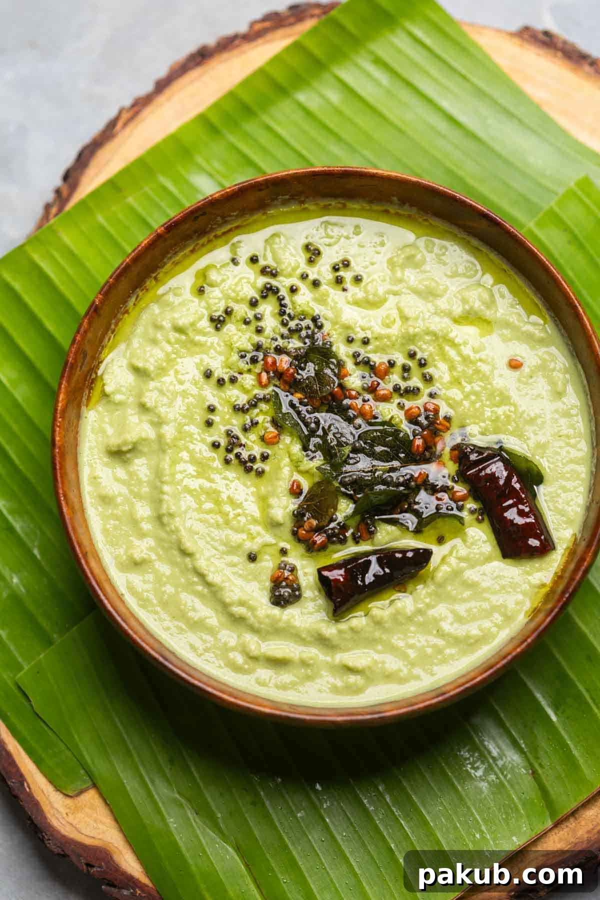 Two pristine white bowls of coconut chutney, one garnished with a red chili tempering and the other with a green herb tempering, showcasing different variations, both placed on a vibrant banana leaf.