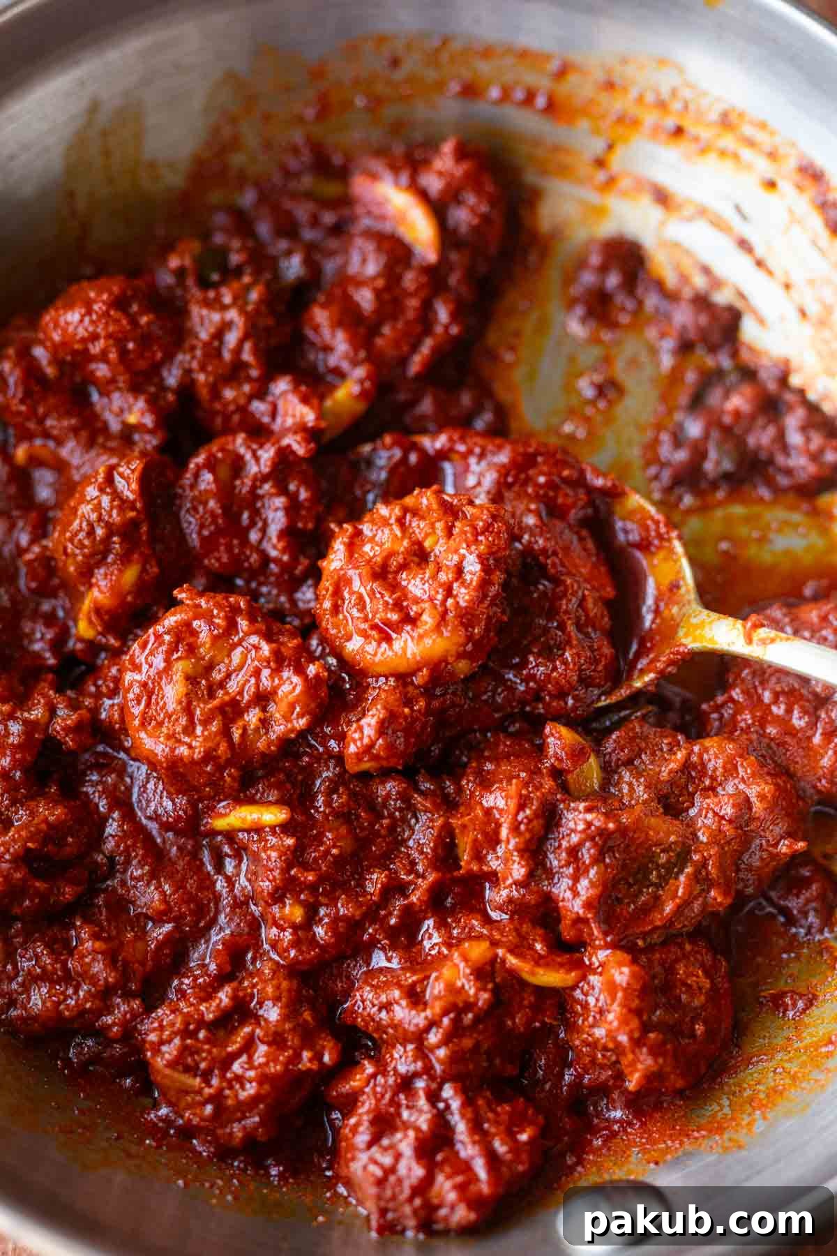 Mangalorean Prawn Pickle in a steel pan with a spoon lifting a prawn pickle up, showcasing its rich, reddish-brown color and whole prawns.