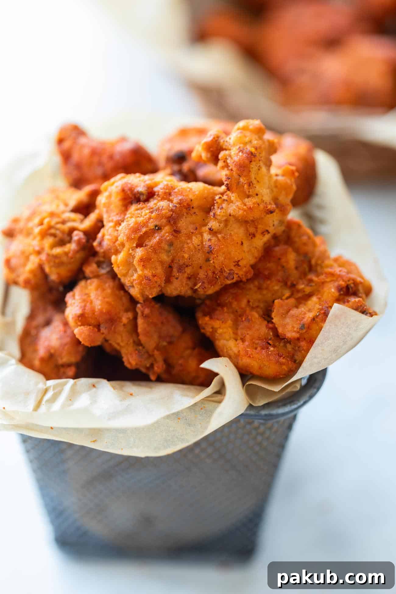 A vibrant close-up of golden-brown KFC style spicy popcorn chicken bites, served in a miniature red and white striped bucket, accompanied by a small bowl of creamy sriracha mayo dipping sauce. The chicken pieces are perfectly breaded, showcasing their crispy texture.